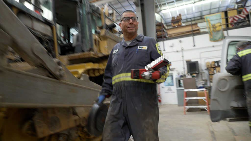 a heavy equipment mechanic in grey coveralls holding power tools